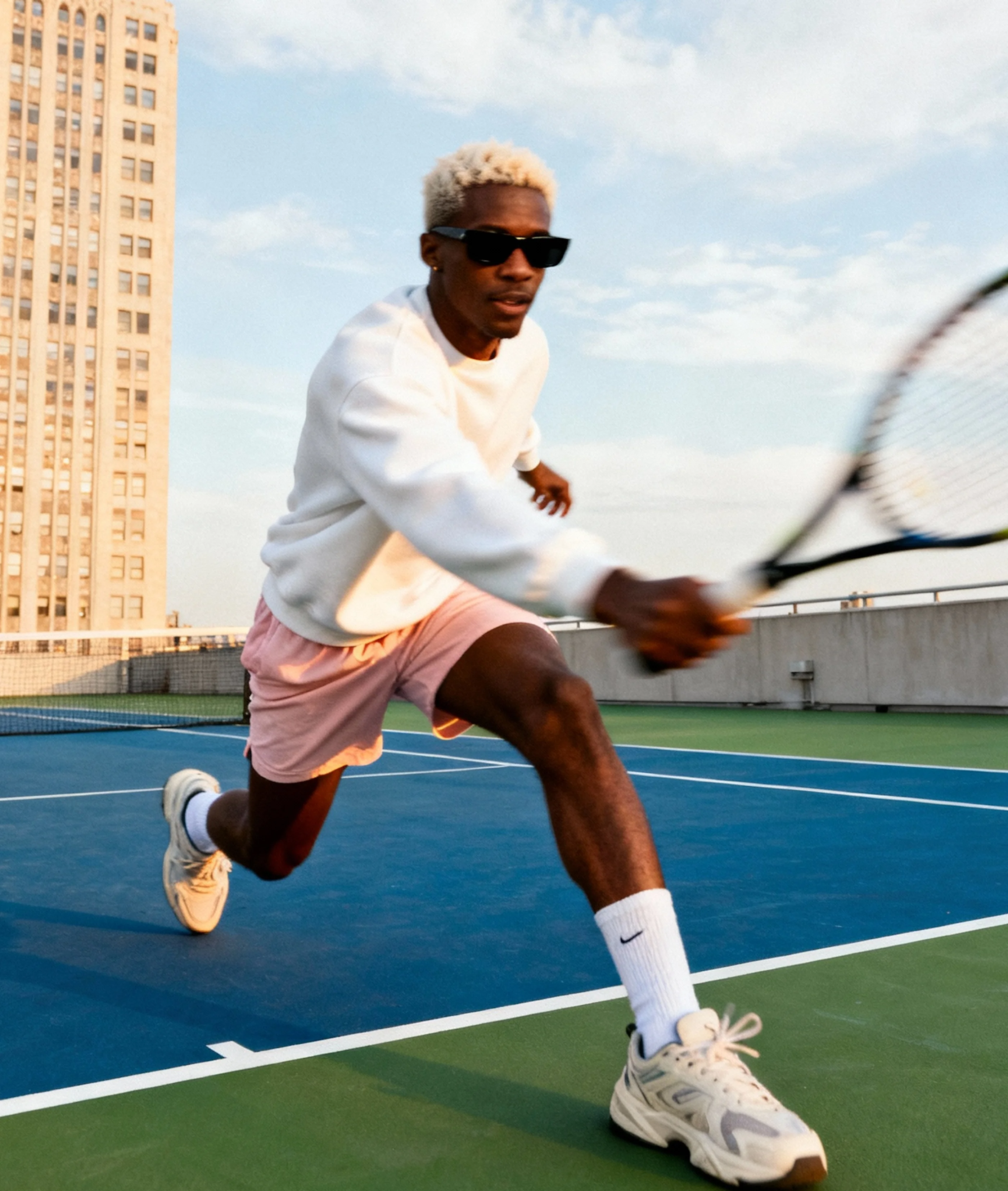 Athletic man in white sweatshirt and pink shorts lunging on an outdoor tennis court while swinging a racket.